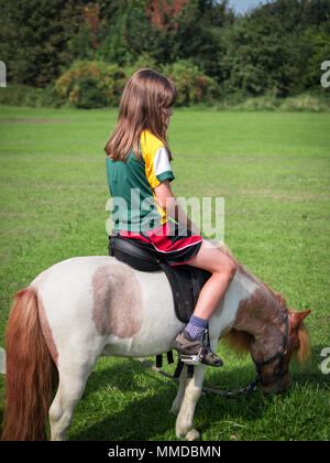 Mädchen sass Reiten auf Miniatur Pferd Beweidung in Feld weg weist (Vertikal) Stockfoto