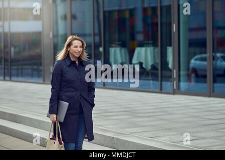 Attraktive stilvolle blonde Frau verlassen Ihren Arbeitsplatz als sie Spaziergänge eine ruhige high key Urban Street lächelnd, als sie ihren Laptop trägt mit Kopie Raum Stockfoto