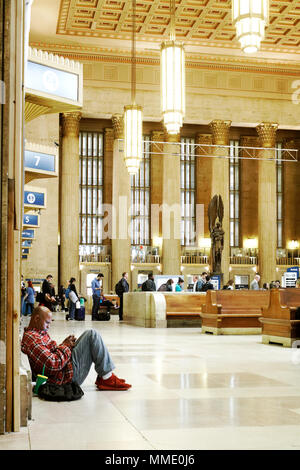 Philadelphia, PA, USA - 23. April 2018: ein Mann sitzt auf dem Boden und verliert sich in sein Handy während der Wartezeit in einem Bahnhof. Stockfoto