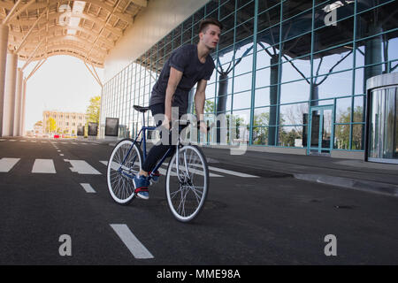 Junger Mann auf dem Fahrrad in der Stadt. Mann auf blauen Fahrrad mit weißen Räder, großer Spiegel Windows Hintergrund Stockfoto