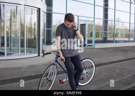 Junge Hipster mit Handy, fixed Gear Bike auf Stadt Straße. großer Spiegel windows Hintergrund, städtische Fahrrad Fahrer Stockfoto