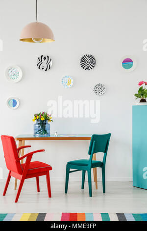 Rot und Grün Stuhl, Platten als Wandschmuck, Lampe, Tisch mit Blumen und bunten Teppich im Wohnzimmer Innenraum Stockfoto