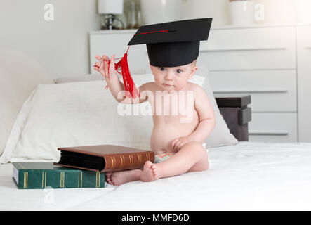 Portrait von Smart baby boy in mortarboard Kappe sitzt und Bücher im Schlafzimmer Stockfoto