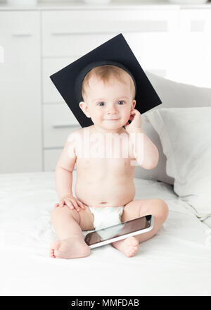 Portrait of smiling baby boy in Windeln tragen Universität Skalenhaube saß auf dem Bett und die Verwendung von Tablet-PC Stockfoto