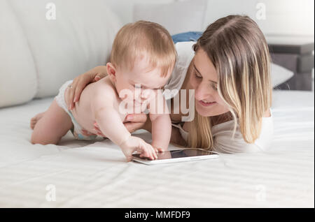 Cute Baby boy in Windeln liegend auf Bett mit Mutter und Verwendung digitaler Tablet Stockfoto