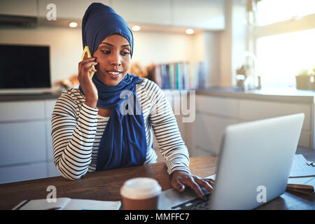 Junge muslimische Frauen Unternehmer trägt ein Kopftuch, an einem Tisch in ihrer Küche sprechen über ein Handy und ein Laptop Stockfoto