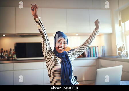 Junge muslimische Frauen Unternehmer Tragen eines hijab heben ihre Arme in Erfolg, während in ihrer Küche, die an einem Notebook arbeitet begeistert Stockfoto