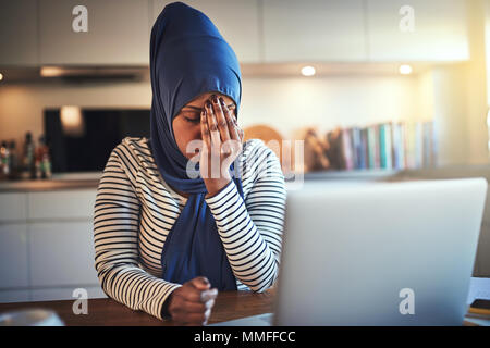 Die junge arabische weibliche Unternehmer Tragen eines hijab an ihrem Küchentisch sitzen mit einem Laptop von zu Hause aus arbeiten Betonte Stockfoto