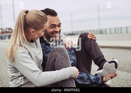 Diverse junge Paar sitzt auf der Straße und zu lachen, während sie für einen Durchlauf zusammen an einem bewölkten Tag Stockfoto