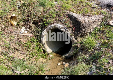 Beschädigte Beton Abflussrohr Ausfahrt mit fließendem Wasser und mit Schmutz, Stein, ungeschnittenem Gras umgeben, getrocknete Blätter und kleine beschädigt Kunststoffrohr Stockfoto