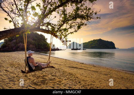 Junger Mann genießt den Sonnenuntergang auf einer Schaukel an einem Strand in Thailand. Stockfoto