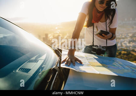 Junge Alleinreisende auf Handy und Touristische Karte auf Auto Motorhaube schauen. Junge weibliche Touristen auf einer Reise auf der Suche nach Richtungen. Stockfoto