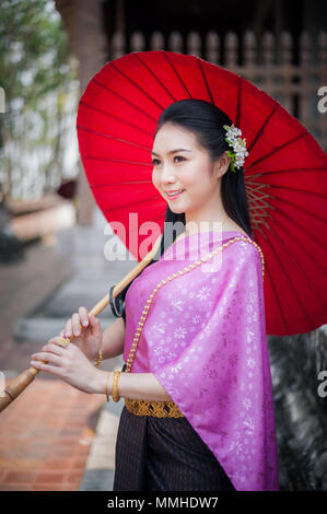 Porträt der schönen thailändischen Frau in Thai national Dress an der Tempel mit roten Regenschirm. Sie lächelt und schaut in die Kamera. Stockfoto