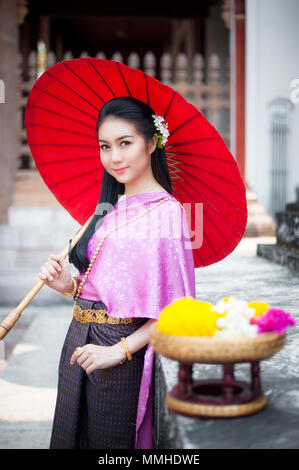 Porträt der schönen thailändischen Frau in Thai national Dress an der Tempel mit roten Regenschirm. Sie lächelt und schaut in die Kamera. Stockfoto