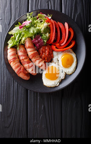 Hausgemachte gebratene Würstchen, Speck, Eier, Soße und Salat in der Nähe verpackt - auf einer Platte. Vertikal oben Ansicht von oben Stockfoto