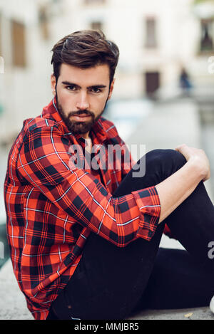 Junger bärtiger Mann, Modell der Mode, trug einen karierten Hemd mit Sitzen in der Straße. Kerl mit Bart und moderne Frisur im städtischen Hintergrund. Stockfoto