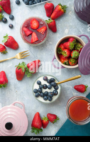 Blick von oben auf die Erdbeeren und Blaubeeren edle Käsekuchen in Glas Glas über einen grauen Hintergrund, mit farbigen Cocotte in Blaubeeren, strawberri Stockfoto