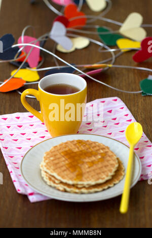 Runde Wafer, gelbe Tasse mit einem langen Löffel auf eine Serviette, mit einem Herzen aus Papier im Hintergrund Stockfoto