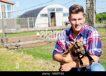 Schöne blonde Mann hält ein Baby Ziege vor einer Scheune Stockfoto