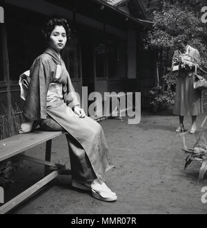 1950, historische, attraktive junge erwachsene Frau sitzen auf einer Bank ausserhalb das Tragen der traditionellen japanischen Kleidung oder Kostüm, den Kimono und durch eine westliche Dame fotografiert werden. Der Kimono ist eine Seide Mantel, um den Körper gewickelt ist, mit einem breiten Gürtel, eine Obi-gebunden und ist die nationale Kleidung für Japan. Stockfoto