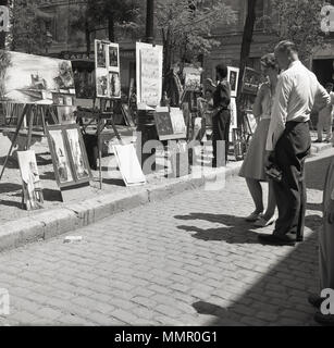 1950, Paar stehend an das Kunstwerk auf der Suche angezeigt auf dem Platz des Place du Tertre am Montmartre, Paris, Frankreich. Das Gebiet, im nördlichen Teil der Stadt, ist berühmt für seine künstlerische Geschichte und Künstler auf der Straße mit den Hauptplatz zu erstellen und Ihre Arbeit. Stockfoto