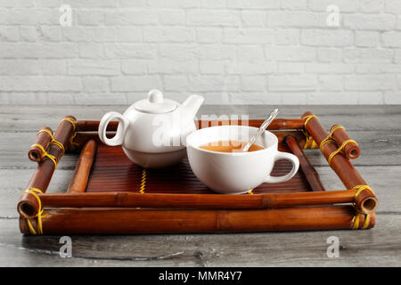 Kleine weiße Keramik Teekanne und Tasse heißen dampfenden schwarzer Tee mit silbernen Löffel serviert auf alte Bambus Fach auf grau Holz Schreibtisch platziert. Stockfoto