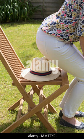 Frau über auf einem Strohhut auf einer hölzernen Stuhl in einem Land Garten positioniert sitzen. Stockfoto