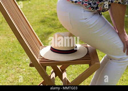 Frau über auf einem Strohhut auf einer hölzernen Stuhl in einem Land Garten positioniert sitzen. Stockfoto