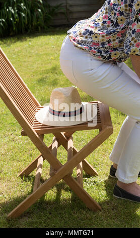 Frau über auf einem Strohhut auf einer hölzernen Stuhl in einem Land Garten positioniert sitzen. Stockfoto