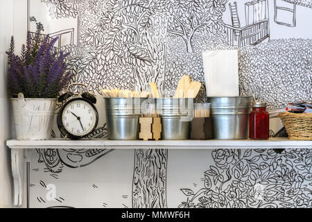 Weiß Regal mit Blumen in einer Ladeschaufel, mit Wecker und verschiedenen Küche Zubehör an der weißen Wand Hintergrund mit Muster. Stockfoto