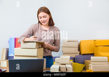 Jungen asiatischen online Verkäufer Geschäftsfrau, die von zu Hause aus arbeiten schreiben Sie den Namen der Kunden auf Ihrem Notizblock und durch Boxen ihrer Produkte umgeben, gute Stockfoto
