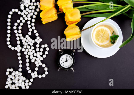 Schwarz Wecker in der Nähe von Bouquet gelbe Tulpen und Schale der Zitrone Tee auf schwarzen Hintergrund. Flach. Mutter oder Frau Tag. Grußkarte. Guten Morgen bre Stockfoto