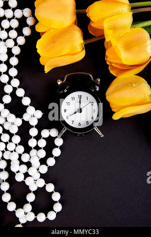 Schwarz Wecker in der Nähe von Bouquet gelbe Tulpen auf schwarzem Hintergrund. Flach. Mutter oder Frau Tag. Grußkarte. Guten Morgen Frühstück. Kopieren Sie Platz. Stockfoto