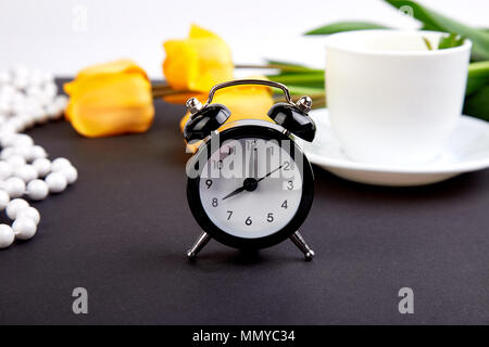 Schwarz Wecker in der Nähe von Bouquet gelbe Tulpen und Schale der Zitrone Tee auf schwarzen Hintergrund. Mutter oder Frau Tag. Grußkarte. Guten Morgen Frühstück. Co Stockfoto