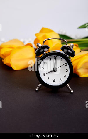 Schwarz Wecker in der Nähe von Bouquet gelbe Tulpen auf schwarzem Hintergrund. Mutter oder Frau Tag. Grußkarte. Guten Morgen. Kopieren Sie Platz. Feder. Stockfoto