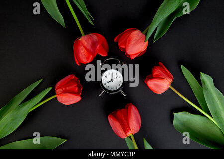 Schwarz Wecker in der Nähe von Bouquet rote Tulpen und Schale der Zitrone Tee auf schwarzen Hintergrund. Mutter oder Frau Tag. Grußkarte. Guten Morgen Frühstück. Kopieren s Stockfoto