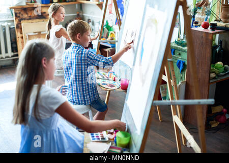 Seitenansicht Portrait von drei Kinder malen auf Staffeleien während der Art Klasse in sonnendurchflutete Studio, auf talentierte junge in der Mitte konzentrieren Stockfoto