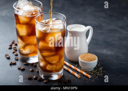 Eis Kaffee in einem großen Glas über und Kaffeebohnen auf einem alten rustikalen Holztisch. Kalten Sommer trinken auf einem dunklen Hintergrund mit kopieren. Der Prozess der Gießen trinken aus einer Kaffeekanne in ein Glas Stockfoto