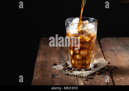 Eis Kaffee in einem großen Glas über und Kaffeebohnen auf einem alten rustikalen Holztisch. Kalten Sommer trinken auf einem dunklen Hintergrund mit kopieren. Der Prozess der Gießen trinken aus einer Kaffeekanne in ein Glas Stockfoto