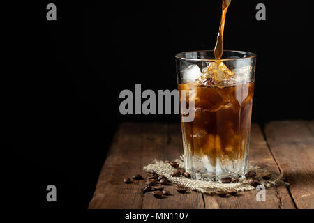 Eis Kaffee in einem großen Glas über und Kaffeebohnen auf einem alten rustikalen Holztisch. Kalten Sommer trinken auf einem dunklen Hintergrund mit kopieren. Der Prozess der Gießen trinken aus einer Kaffeekanne in ein Glas Stockfoto