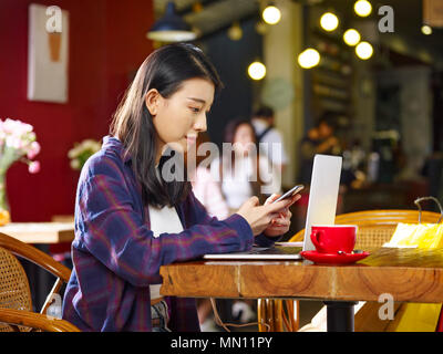 Junge asiatische Frau sitzt im Café mit Handy und Laptop. Stockfoto