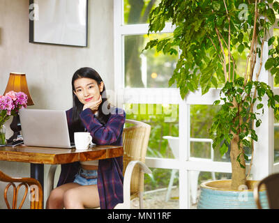 Schöne junge asiatische Frau an der Kamera schaut lächelnd, während sie Laptop im Coffee Shop. Stockfoto