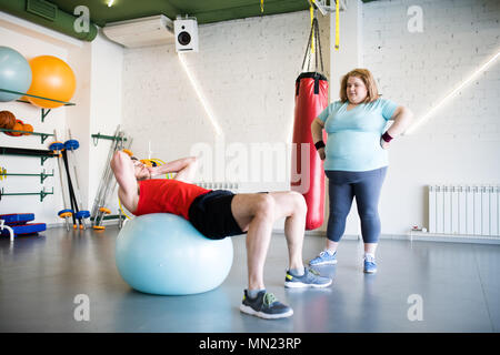 In voller Länge Porträt der übergewichtigen Jungen Frau beobachten, stattlicher Mann Übungen auf Fitness Ball in der Turnhalle, kopieren Raum Stockfoto