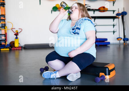 Volle Länge Portrait von erschöpft beleibte Frau sitzt auf Schritt und Trinkwasser nach extremen weightloss Workout im Fitness Club, Platz kopieren Stockfoto