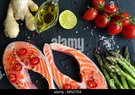 Zwei frischen, rohen Lachs Steaks mit Gemüse und Gewürze: Spargel, Tomaten, Ingwer, Pfefferkörner, Salz, Pfeffer, Chili, Zitronensaft und Olivenöl auf Schwarz ba Stockfoto