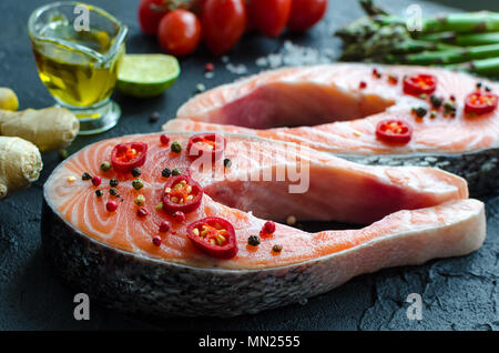 Zwei frischen, rohen Lachs Steaks mit Gemüse und Gewürze: Spargel, Tomaten, Ingwer, Pfefferkörner, Salz, Pfeffer, Chili, Zitronensaft und Olivenöl auf Schwarz ba Stockfoto