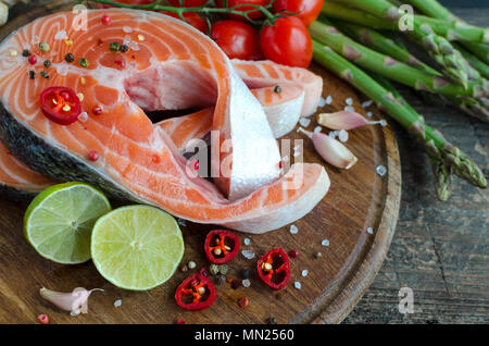 Zwei frash roher Lachs Steaks mit Gemüse und Gewürze: Spargel, Knoblauch, Tomaten, Pfefferkörner, Salz, Chili und Zitrone auf Holz Schneidebrett. Heilen Stockfoto