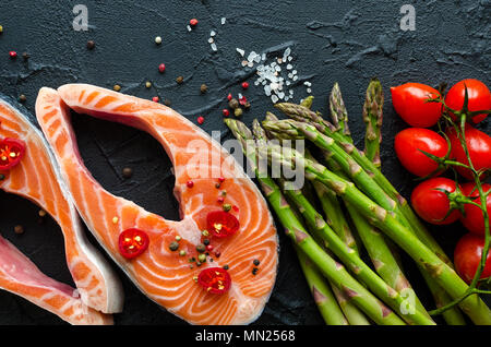 Zwei frischen, rohen Lachs Steaks mit Gemüse und Gewürze: Spargel, Tomaten, Pfefferkörner, Salz, Pfeffer, Chili, Zitronensaft und Olivenöl auf schwarzem Hintergrund Stockfoto