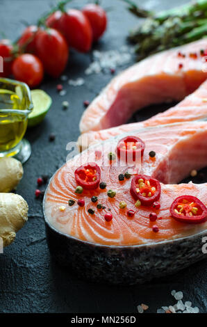 Zwei frischen, rohen Lachs Steaks mit Gemüse und Gewürze: Spargel, Tomaten, Ingwer, Pfefferkörner, Salz, Pfeffer, Chili, Zitronensaft und Olivenöl auf Schwarz ba Stockfoto