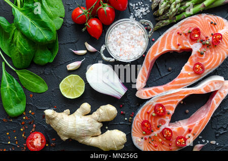 Zwei frischen, rohen Lachs Steaks mit Gemüse und Gewürze: Spargel, Spinat, Tomaten, Knoblauch, Ingwer, Pfeffer, Salz, Chili, Zwiebeln und Zitronen auf Schwarz ba Stockfoto